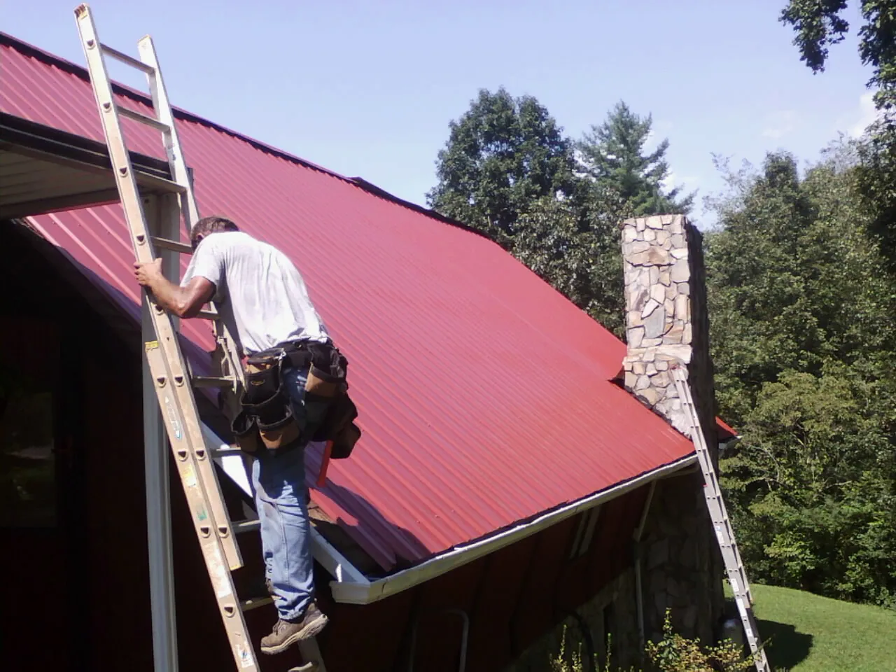 Roofer on a ladder beside a red metal roof and stone chimney — Soffit & Fascia Repair by Roofing Brothers in Felt